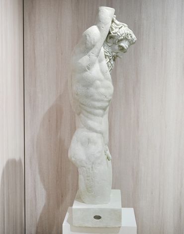 Side view of a classical marble male torso statue with missing arms and head.