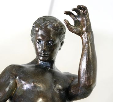 Close-up of the face, raised hand, and upper body of Athlete, Male Nude, a bronze-toned plaster scul