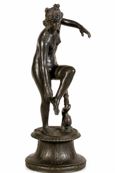 Bronze statue of a woman adjusting her sandal, standing on a decorated base.