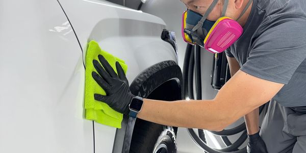 Person cleaning a white car with a yellow cloth and wearing protective gear.