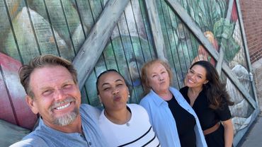 Four people smiling and posing in front of a colorful mural on a wooden fence.