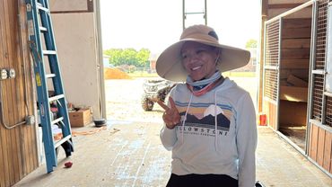 Woman in a wide-brimmed hat making a peace sign inside a barn.
