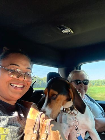 Two women and a dog smiling inside a car during a sunny day.