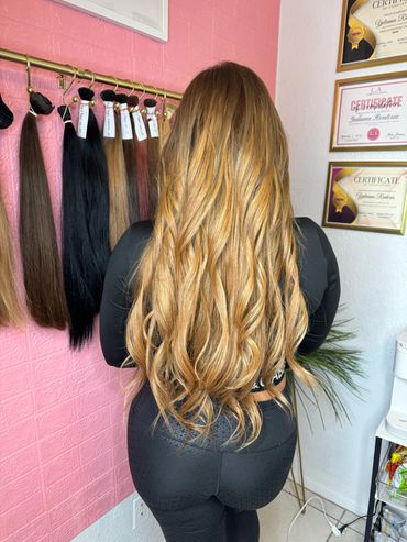 Woman with long, wavy blonde hair standing in a salon with hair extensions displayed.