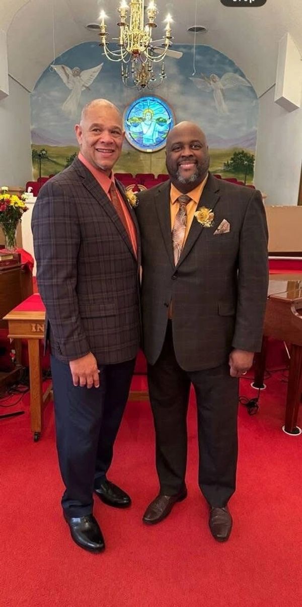 Two men in suits smiling inside a church with a chandelier and stained glass window.