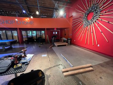Two people assembling wooden platform inside a workshop with tools and construction materials.