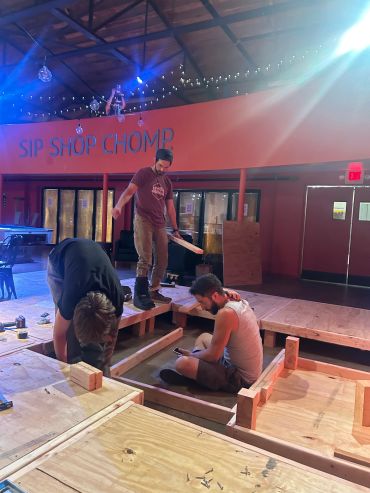 Three men building a wooden structure inside a room with 'SIP SHOP CHOMP' sign.