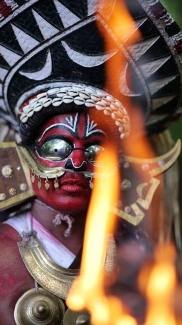 A traditional Theyyam performer in elaborate face paint and costume behind flames.