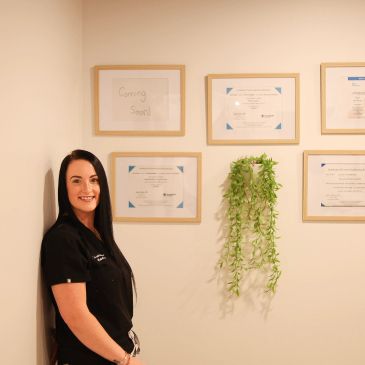 Woman smiling by wall with framed certificates and green plant.