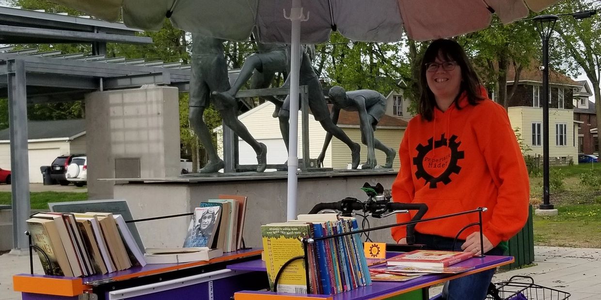 A smiling Book Lady sits on her book bike which is open, displaying books for people to take. 