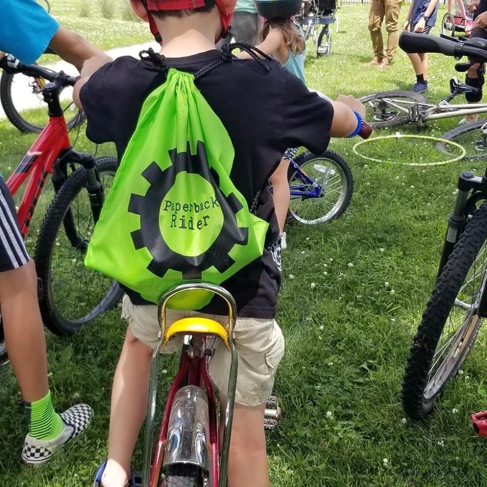 A green string bag with the Paperback Rider logo is on the back of a young boy sitting on his bicycl
