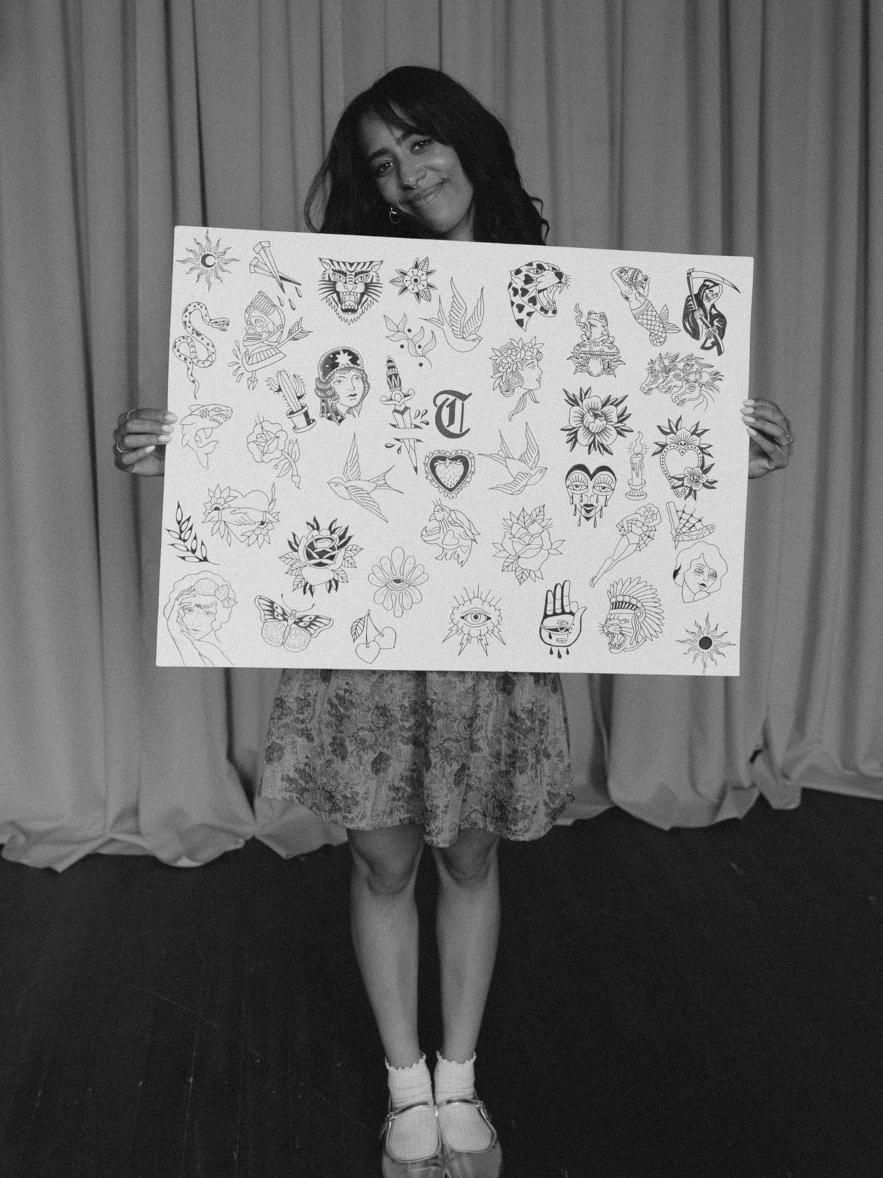 A woman holding a large sheet with various black ink tattoo designs in front of an orange curtain.