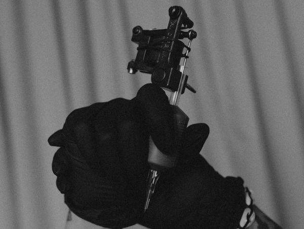 Hands in black gloves holding a tattoo machine against an orange backdrop.