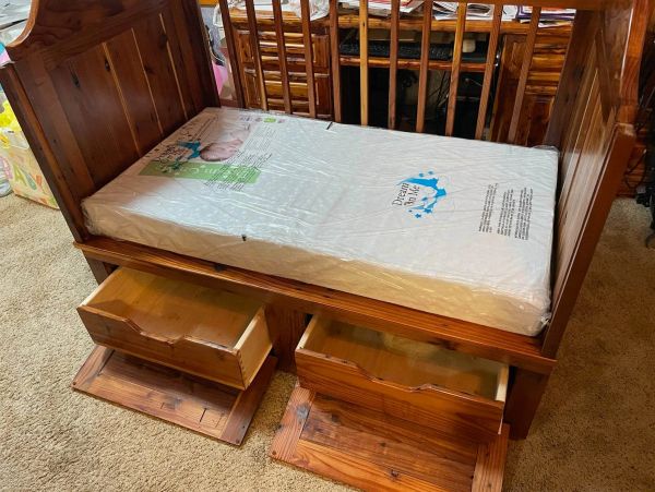 Wooden crib with mattress and two open drawers beneath.