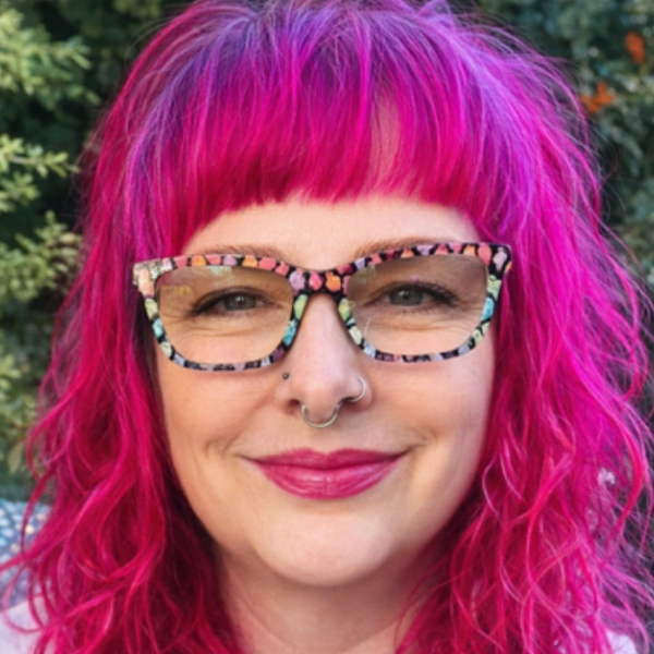 Woman with vibrant pink hair and colorful glasses smiling outdoors.