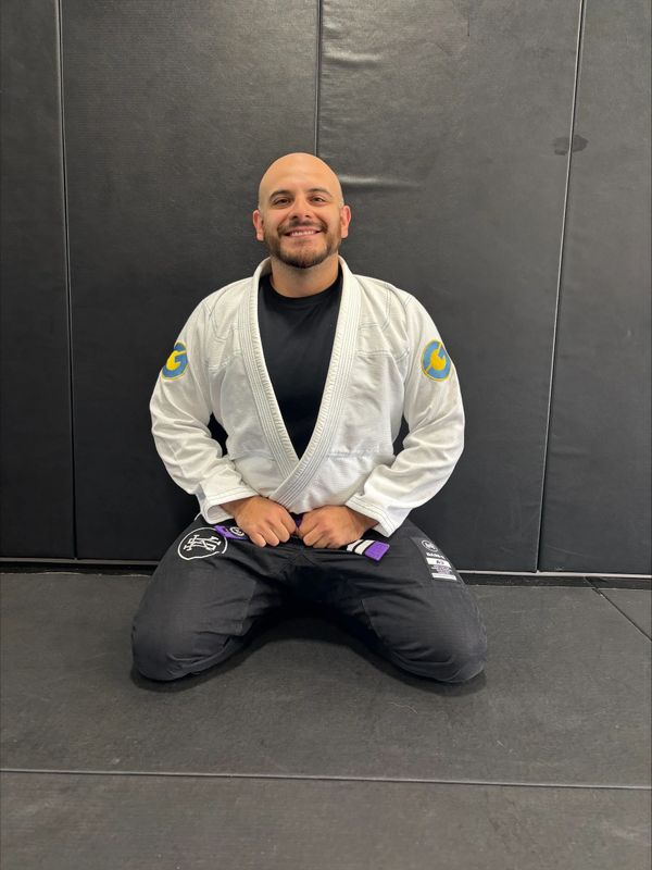 Man in a white Brazilian Jiu-Jitsu gi kneeling and smiling on a mat.