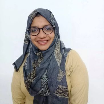 Smiling woman wearing glasses and a patterned grey hijab with a beige top.