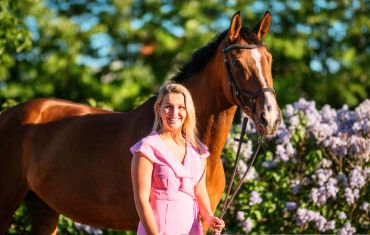 A smiling woman in a pink dress stands beside a brown horse in a lush, sunny garden.