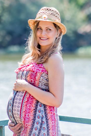 Pregnant woman in a colorful dress and hat smiling outdoors.