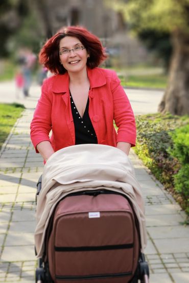 Smiling woman in red jacket pushing a stroller outdoors on a sunny day.