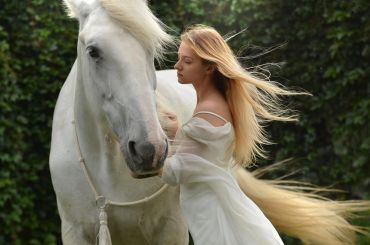 A woman in white gently embraces a white horse with flowing hair.