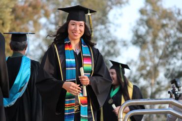 Smiling graduate in cap and gown holding diploma outdoors.