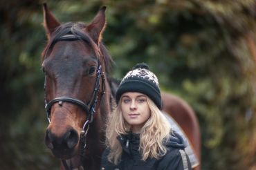 A young woman in a winter hat standing beside a brown horse outdoors.
