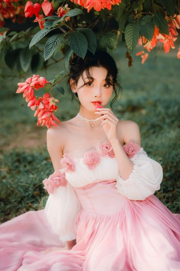 A woman in a pink dress with floral details poses under vibrant red flowers.