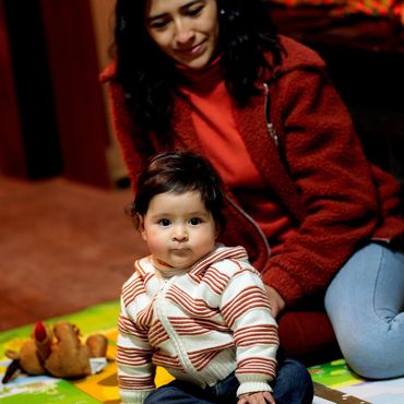 A baby sitting on a colorful mat with a woman smiling behind.
