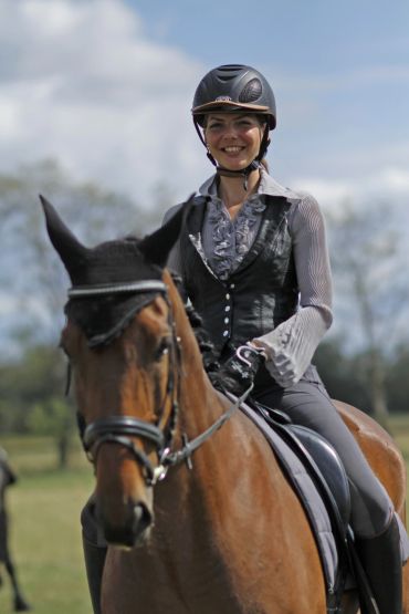 Smiling woman in equestrian gear riding a horse outdoors.
