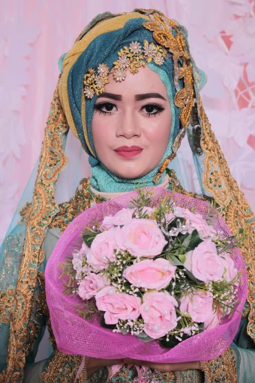 A bride in a detailed blue and gold hijab holding a bouquet of pink roses.