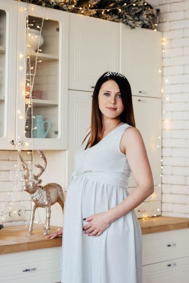 Pregnant woman in a white dress standing in a decorated kitchen with fairy lights.