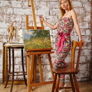 Woman artist posing next to an easel with a landscape painting in a rustic studio.