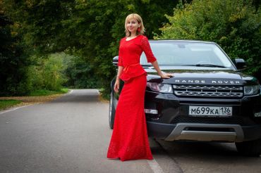 Woman in red dress leaning on a black Range Rover on a forest road.