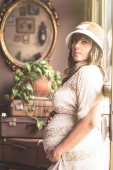 Pregnant woman in a vintage-style room wearing a beige hat and dress.