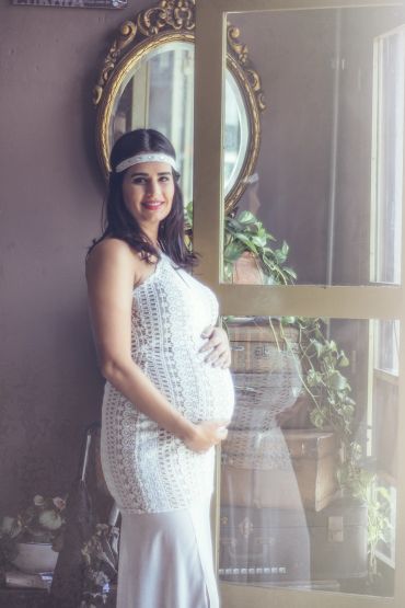 Pregnant woman in a white dress smiles beside a vintage mirror and plants.