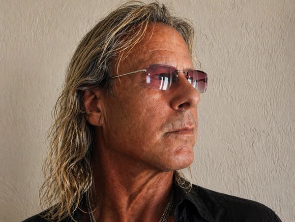 Man with long blond hair, sunglasses, and layered necklaces looks sideways.