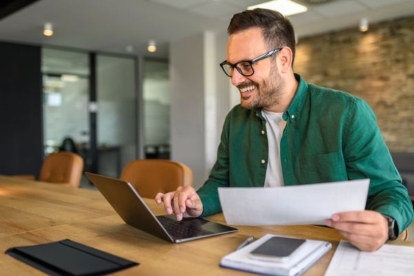 Positive businessman in glasses examining sales report over laptop at desk while working in office