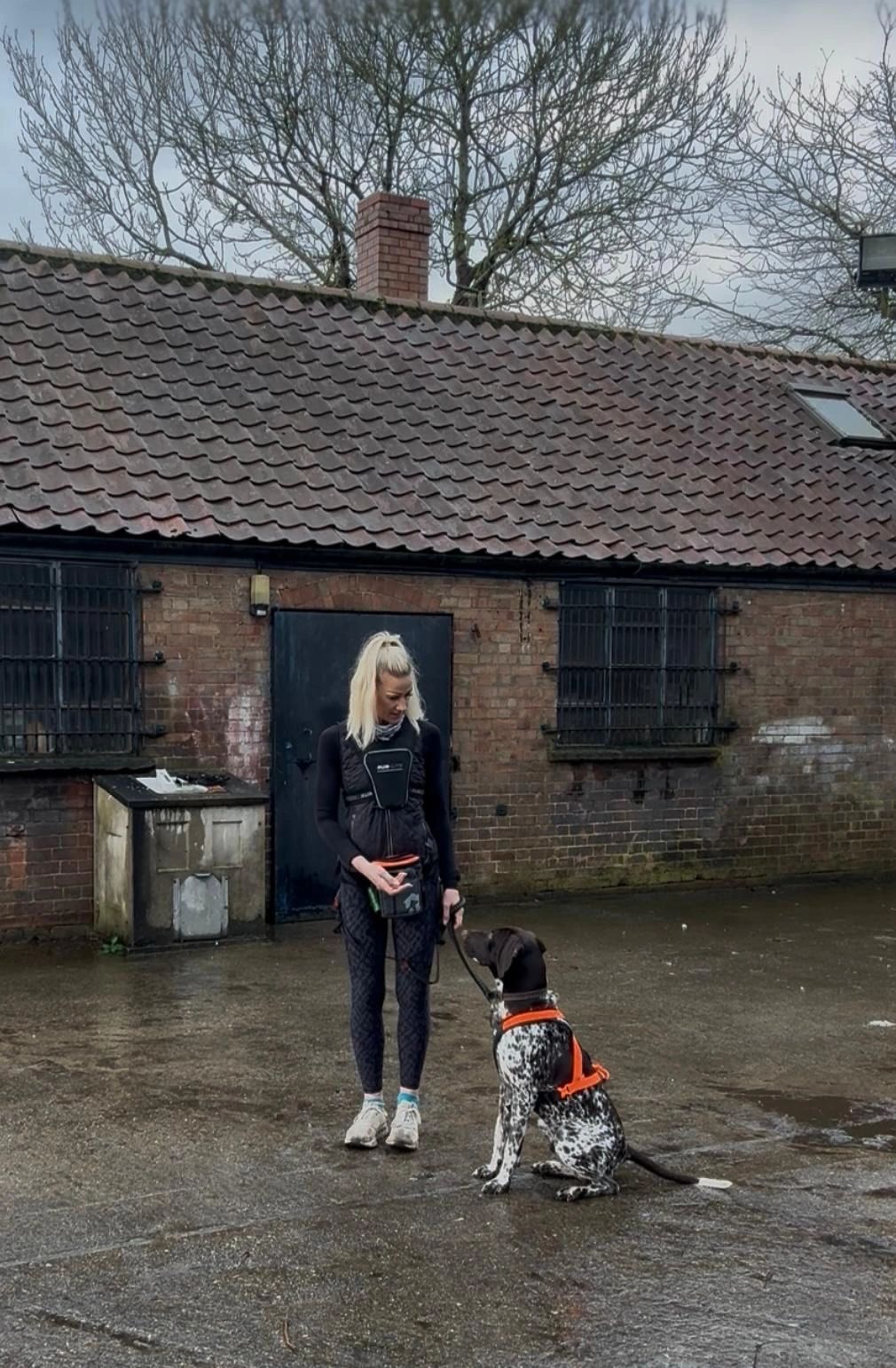 Woman training a dog outside a rustic brick building on a wet day.