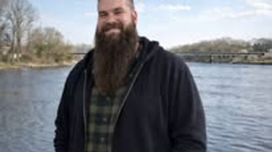 A bearded man smiling near a river on a clear day.