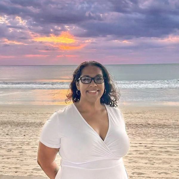 A beautiful woman smiling in front of a beach with the sunset in the background.