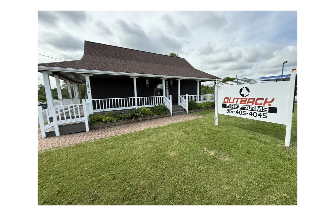 A rustic building with a large sign for Outback Firearms on a grassy lawn.