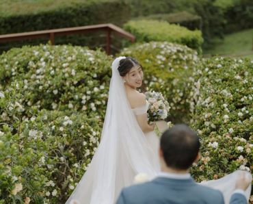 Bride looks back smiling at groom in a lush garden filled with white flowers.