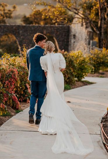 A couple walks together outdoors, the woman in a white wedding dress with a long veil.
