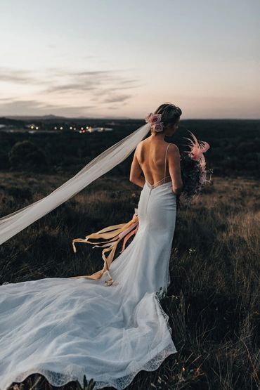Bride in a flowing white dress with a long veil stands in a field at dusk.