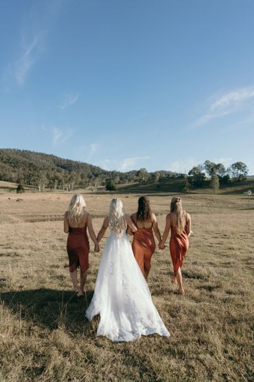 Bride and bridesmaids walking hand in hand in a sunny open field.