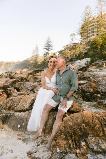Couple sitting on rocks by the beach, sharing a tender moment.
