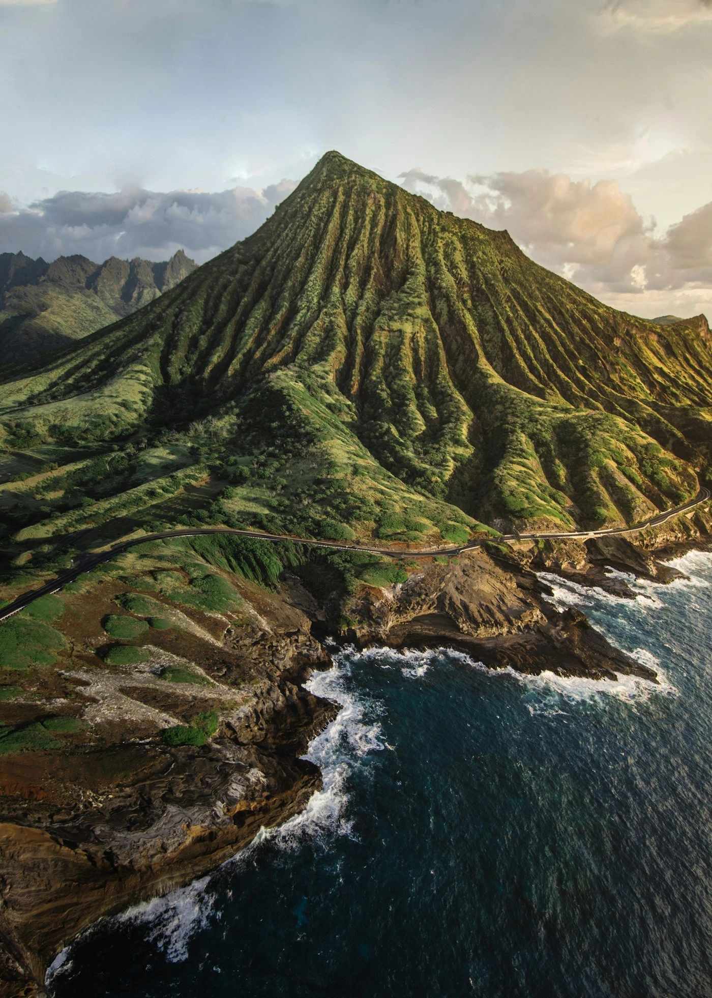 A lush green mountain with deep ridges by the ocean under a cloudy sky.