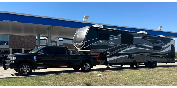 A black truck towing a Montana Legacy RV at a gas station under clear blue skies.