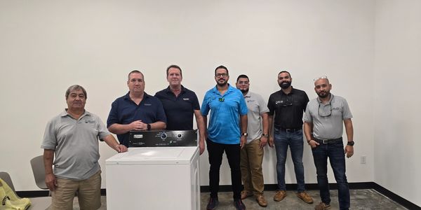 Seven men standing behind a washing machine in a plain room.